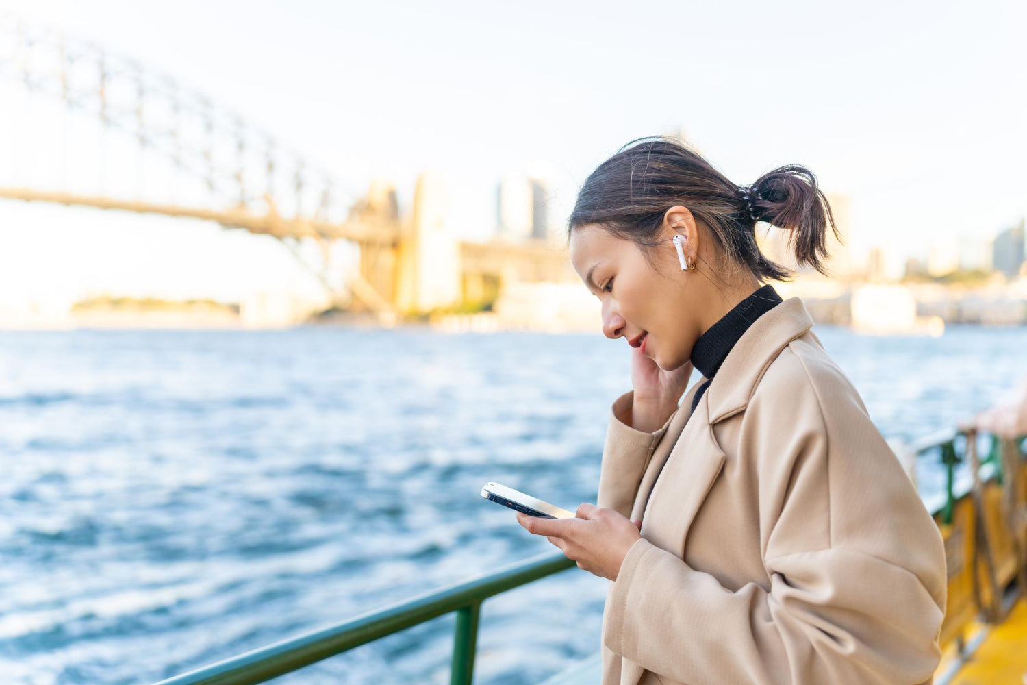 Woman in a beige coat using a smartphone by a waterfront with a bridge in the background